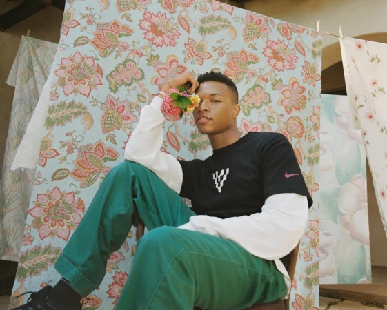 Young Black man holding flowers near patterned fabric hanging on clotheslines by Myles Loftin