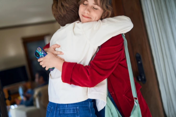 Two people embracing closely, with one smiling woman wearing a red jacket and a light green crossbody bag visible in the frame.