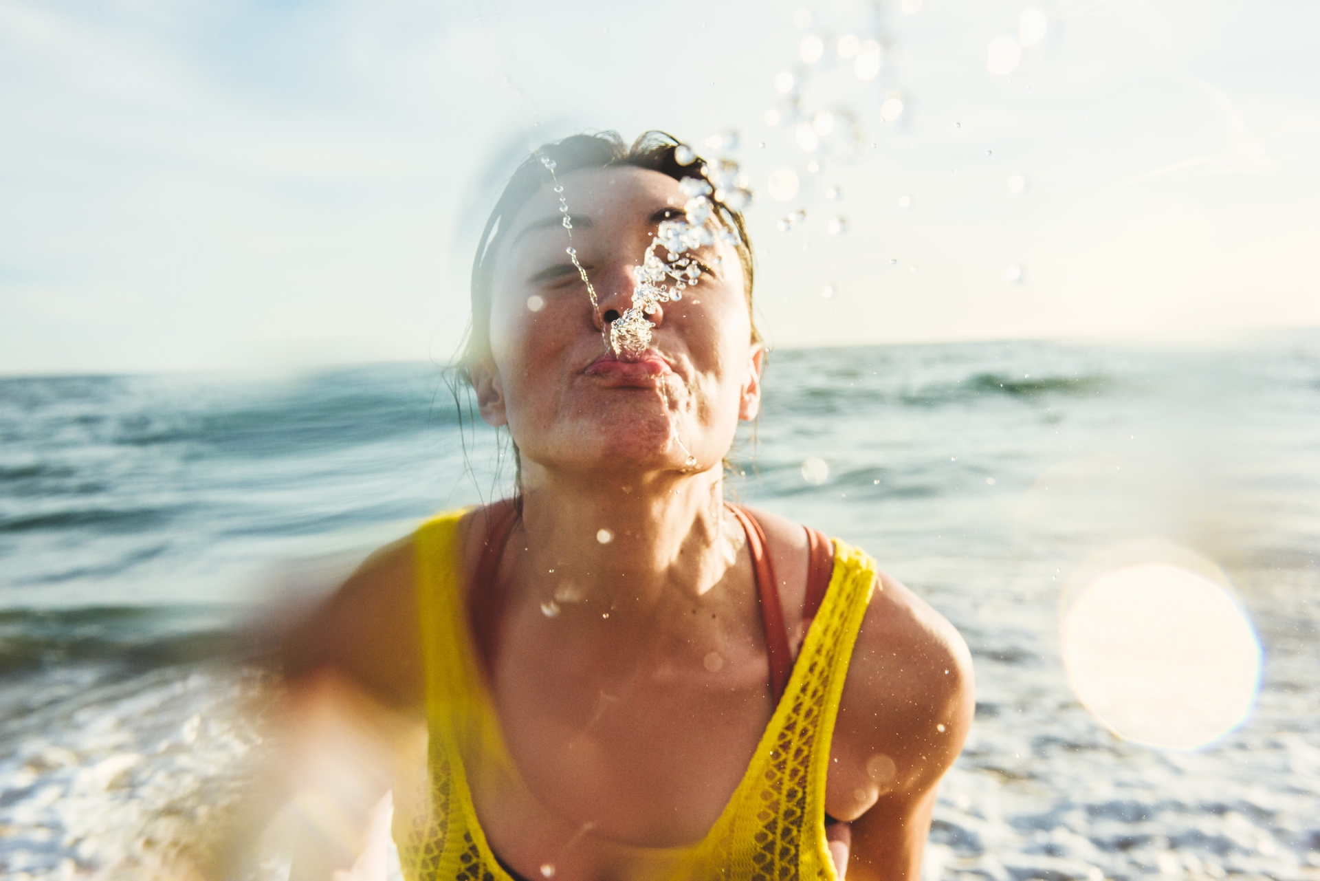 Adult female in a yellow swimsuit spits water into the air on a sunny beach with blue water. Close-up shot with blurred water drops.
