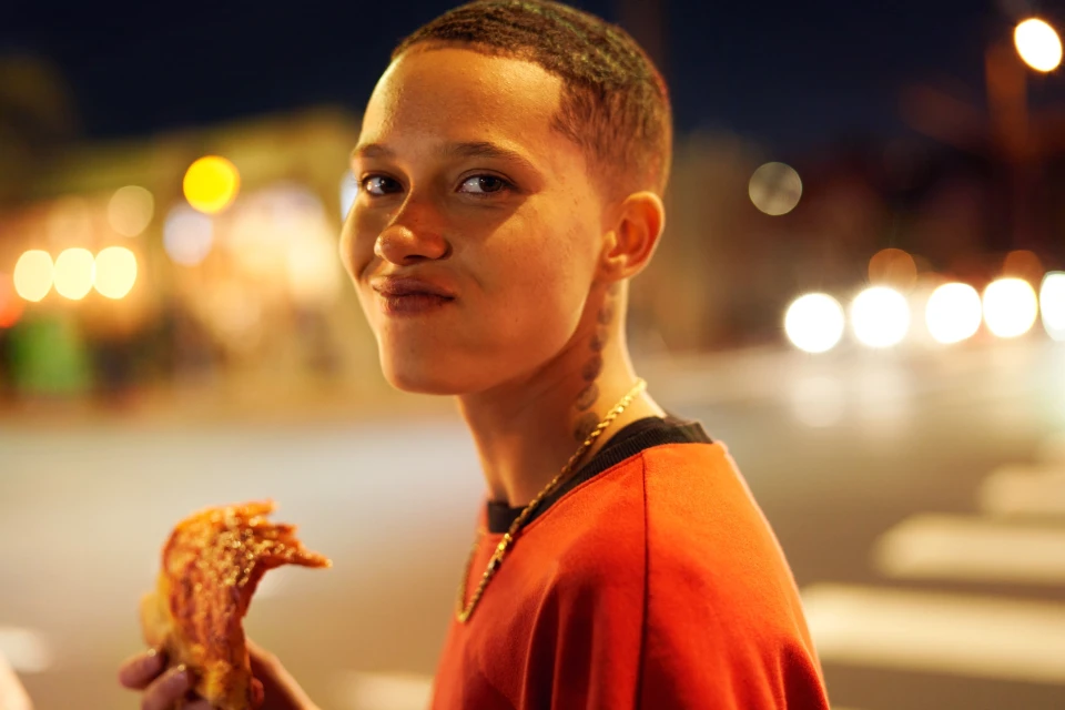 Close-up of a young adult with short hair eating pizza outdoors at night, lit by street lights and smiling at the camera.