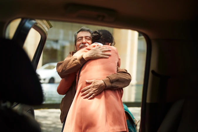 An elderly man and a woman embracing next to a car. The scene is captured from inside the vehicle, showing the warm interaction.