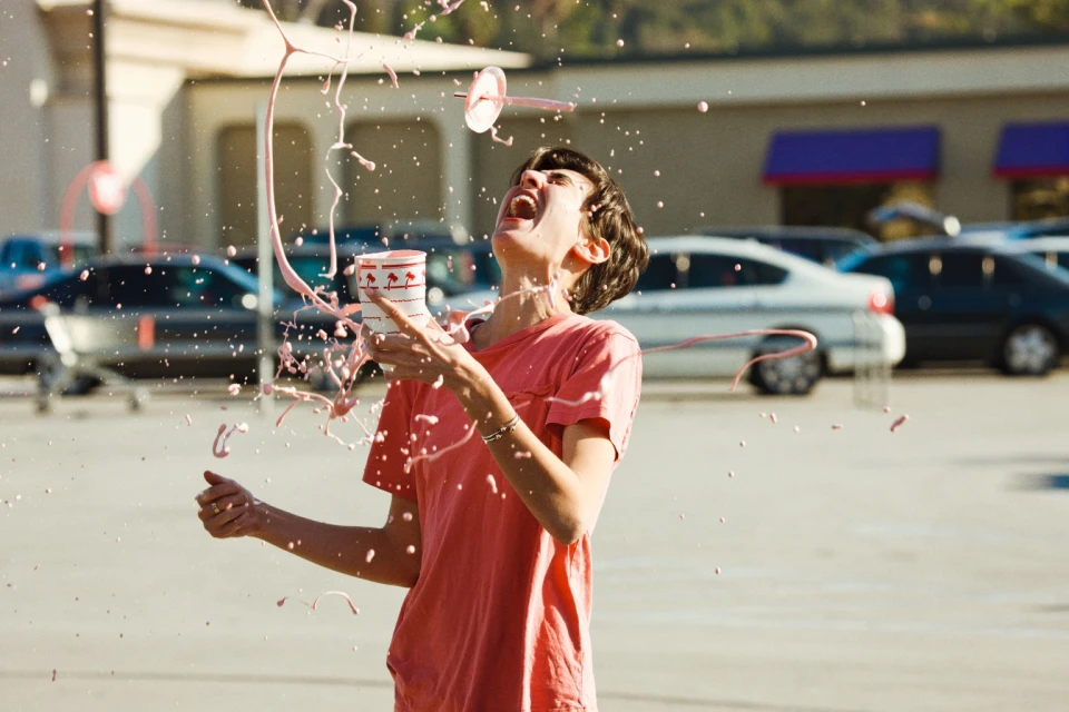 Young adult outdoors in a parking lot, splashed with a pink drink from a cup with a logo on it, cars and a building in the background.