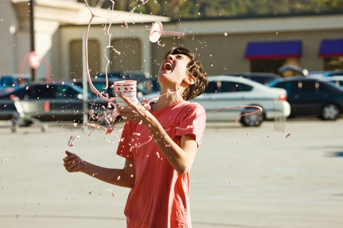 Young adult outdoors in a parking lot, splashed with a pink drink from a cup with a logo on it, cars and a building in the background.