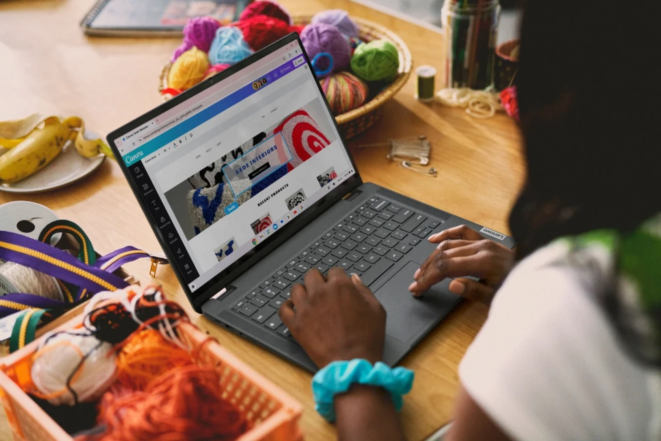 Person using laptop with Canva interface showing designs, surrounded by yarn, ribbon, and sewing items on a wooden table.