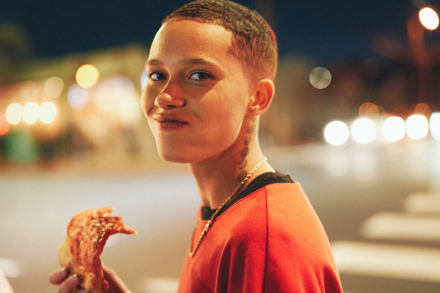 Portrait of a young adult with short hair, eating pizza outdoors at night, illuminated by city lights, wearing a red shirt and gold chain.