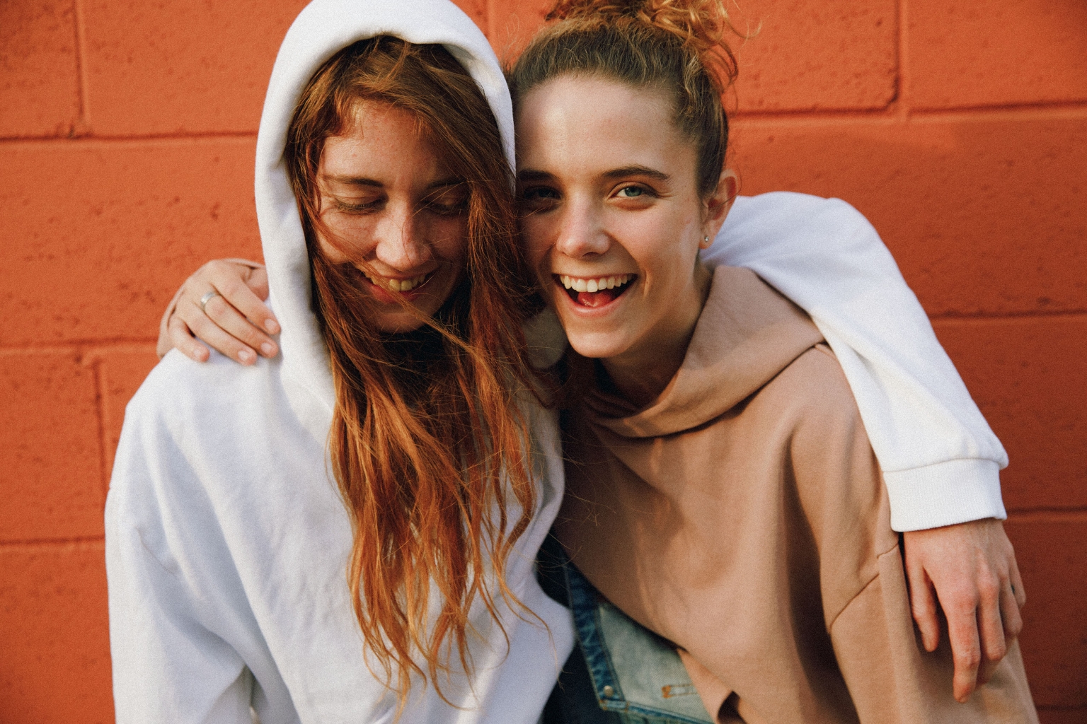 Two cheerful young women embrace outdoors against an orange brick wall, smiling and wearing casual hooded sweatshirts.