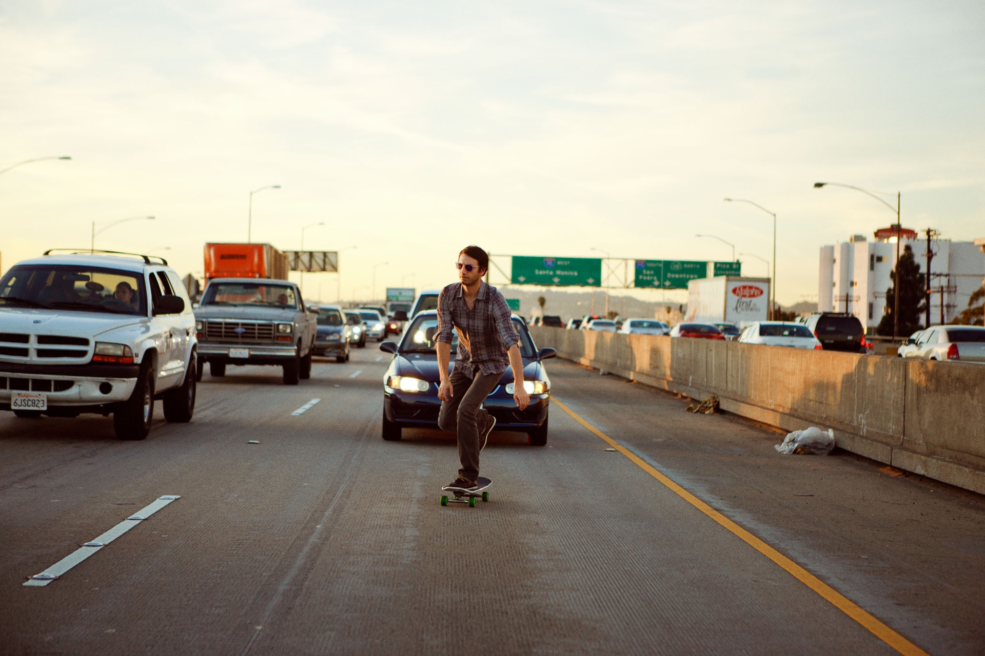 A man is skateboarding on a busy highway with cars in the background during daytime, wearing sunglasses, shirt and pants.