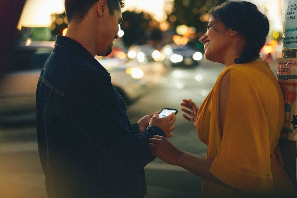 A couple in an urban setting at dusk, with the man holding a smartphone and both smiling; city lights blurred in background.