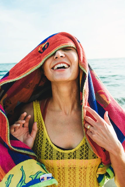 Laughing woman with colorful patterned towel draped over her head stands on a beach with the ocean and sky visible in the background.