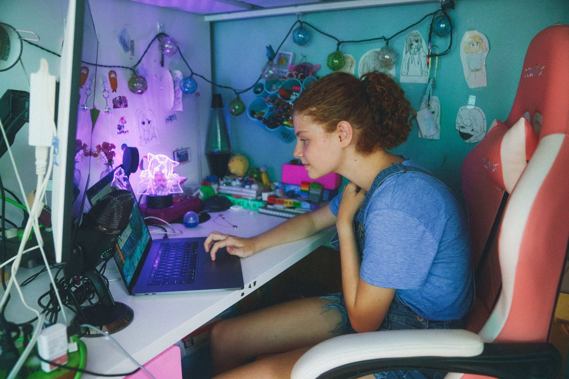 Young woman with curly hair sits at a desk with a laptop, microphone, and monitors, illuminated by blue and purple lighting.