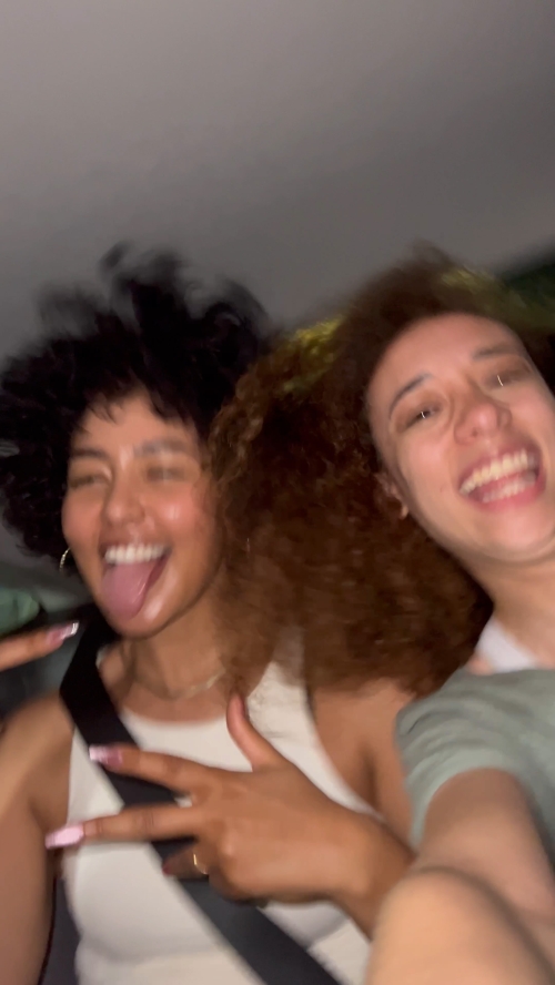 Two smiling women with curly hair pose indoors, making a peace sign with their fingers. The photo has a casual, candid feel.