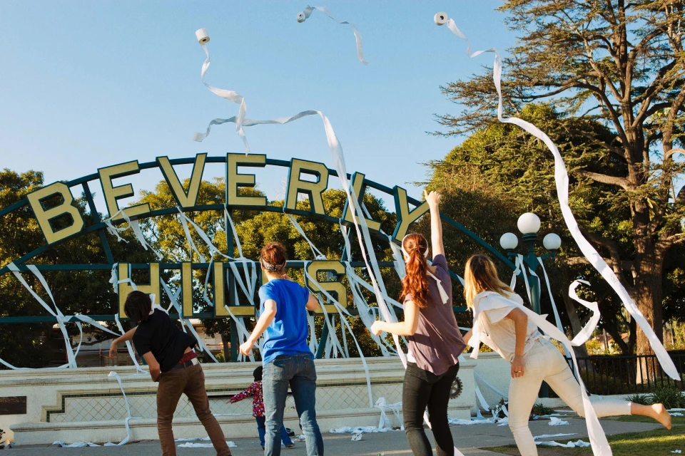 Group of young adults playfully decorating the Beverly Hills sign with toilet paper, on a bright sunny day in an outdoor setting.
