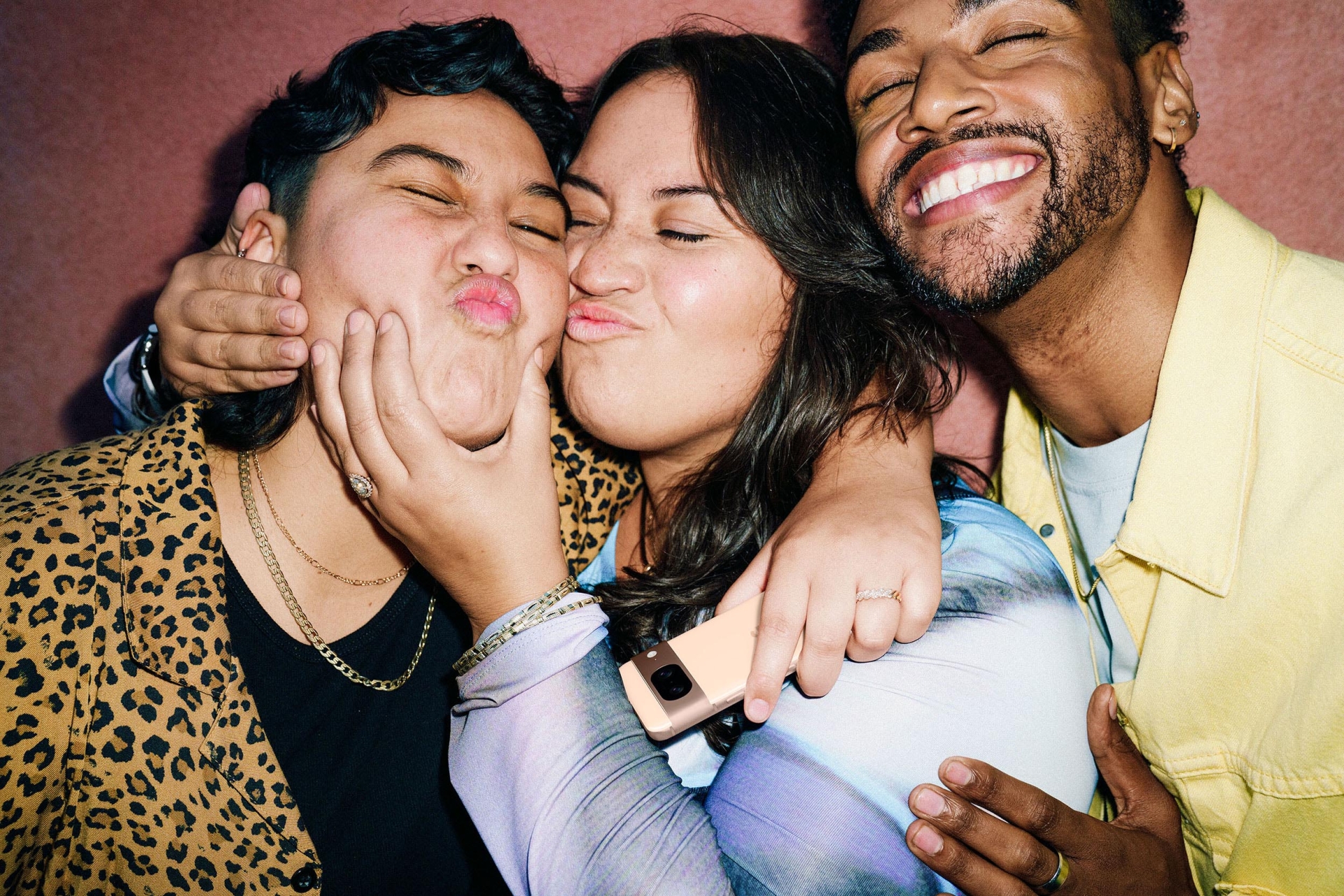 Three cheerful people embrace, making silly faces, posing indoors with a smartphone visible in hand and a pink wall in the background.