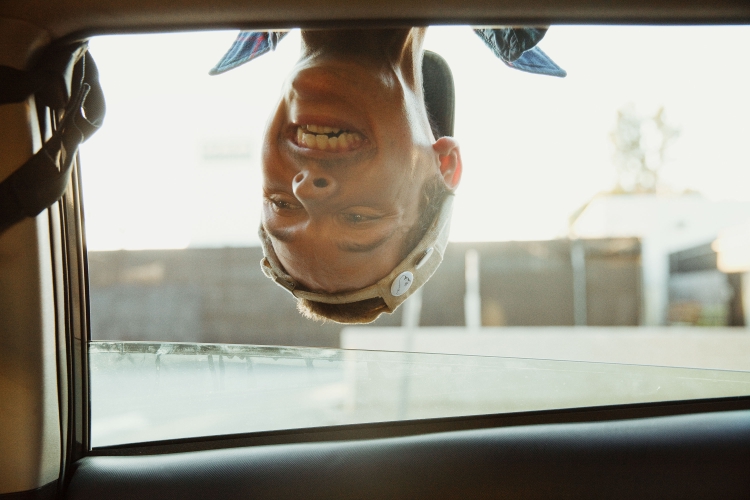 Young adult smiles upside-down, hanging out of a car window, with an urban background visible through the glass on a sunny day.