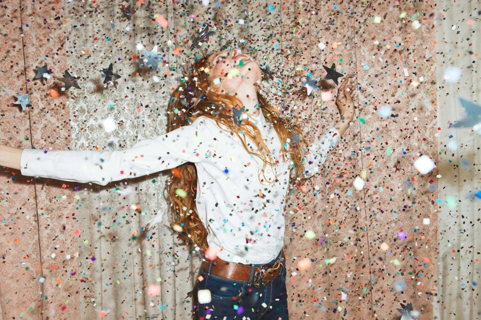 Young adult woman with long red hair poses with outstretched arms as confetti falls in an indoor location with a patterned background.
