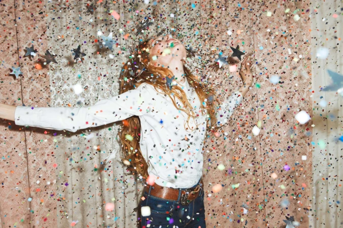 Young adult woman with long red hair poses with outstretched arms as confetti falls in an indoor location with a patterned background.