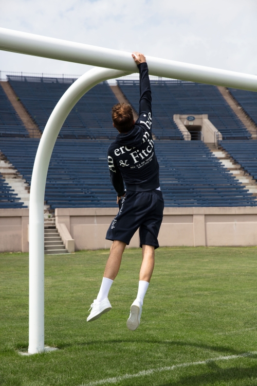 Male athlete wearing casual sporty apparel hanging from a soccer goalpost on a green lawn at an outdoor stadium on a sunny day.