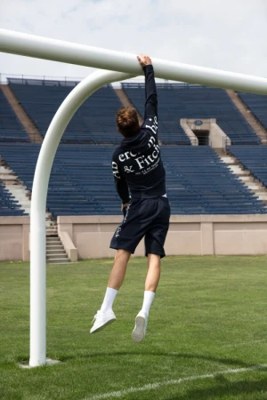 Male athlete wearing casual sporty apparel hanging from a soccer goalpost on a green lawn at an outdoor stadium on a sunny day.