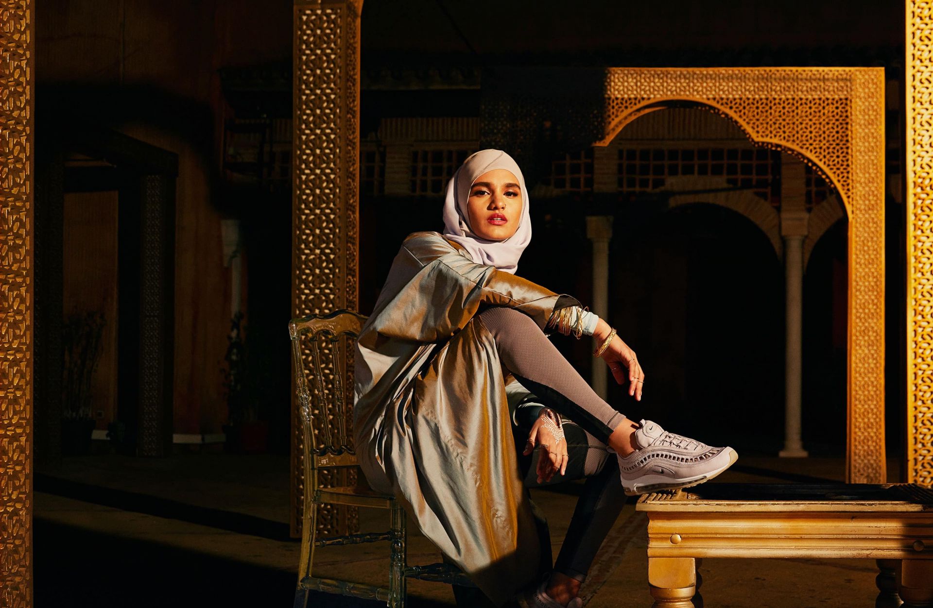 A woman wearing a hijab and athletic shoes sits on an ornate chair in an indoor courtyard with patterned arches and columns.