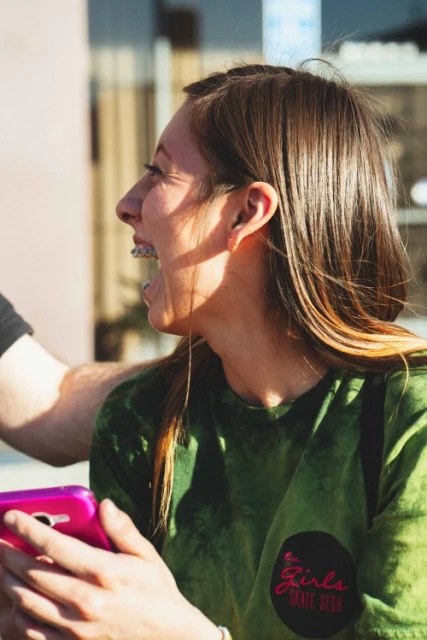 Profile view of a young woman with braces smiling while holding a pink cell phone in an outdoor environment with natural light.