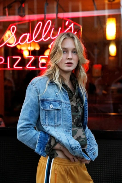 Woman in denim jacket and sweatpants poses in front of a pizza restaurant with neon sign. Urban fashion, outdoor lighting.