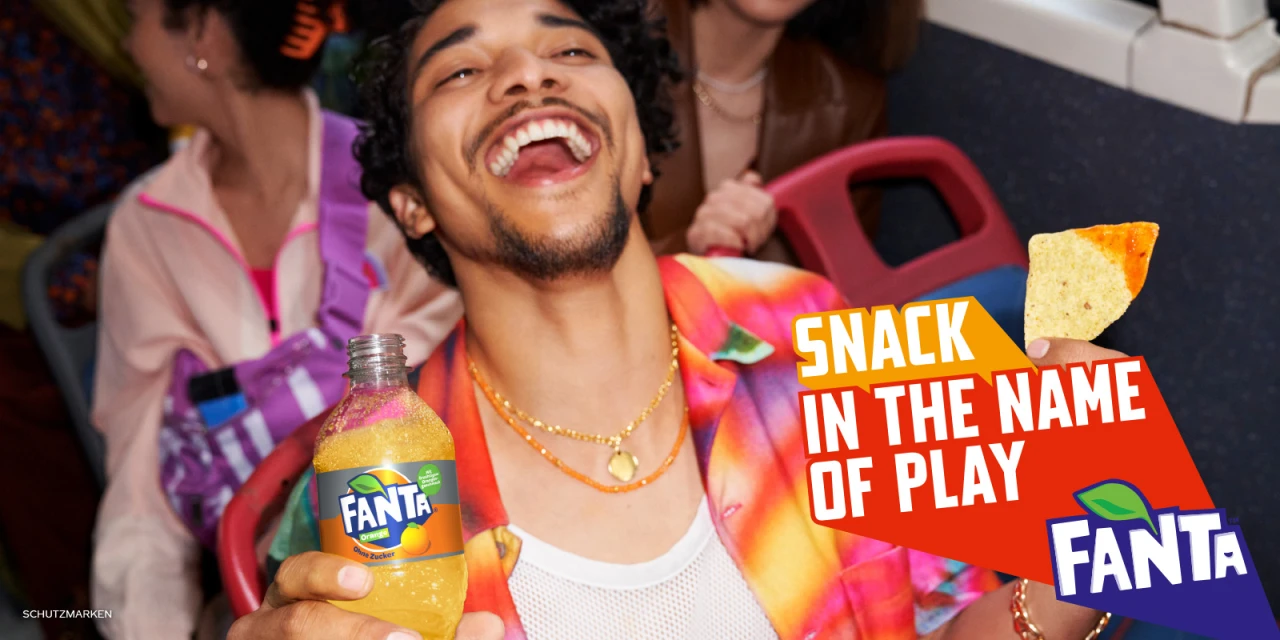 Laughing man in bright shirt holds Fanta in an indoor setting with chips and commercial text: 'Snack in the name of play'.