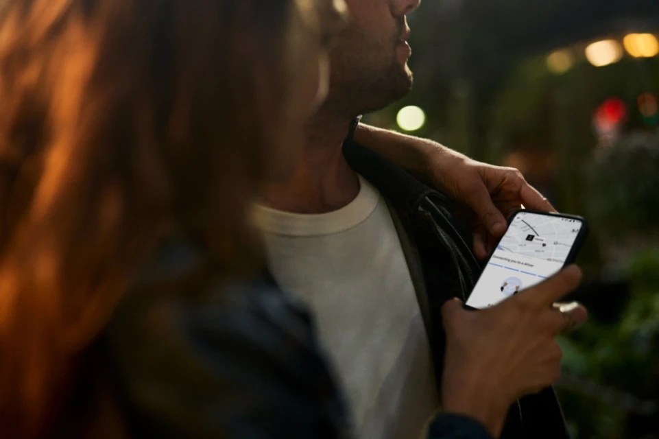 Man and woman on city street at night, woman holds phone with map displayed, illuminated buildings in background. Two people in low light.