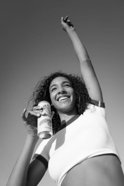 Black and white shot of a woman smiling while holding a can against a bright sky. The woman's arm is raised upward.