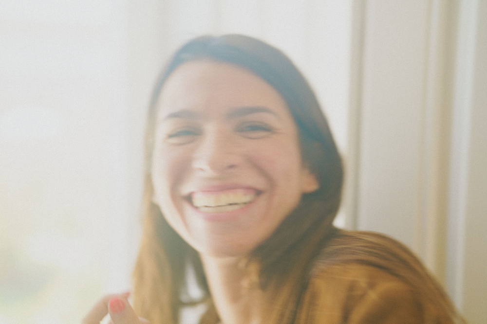 Smiling woman with brown hair indoors near window with natural light, wearing a tan shirt, happy expression, beauty and lifestyle.
