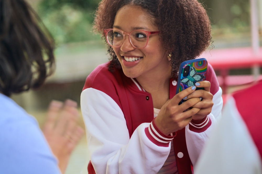 Smiling woman with curly hair holds her phone with a colorful case and is wearing a maroon and white letterman-style jacket indoors.