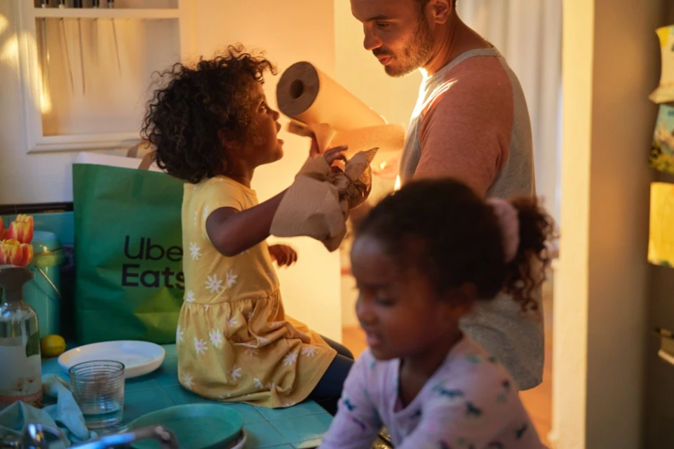 Father and two daughters in kitchen, one child sitting on counter next to an Uber Eats bag, all interacting with brown paper.