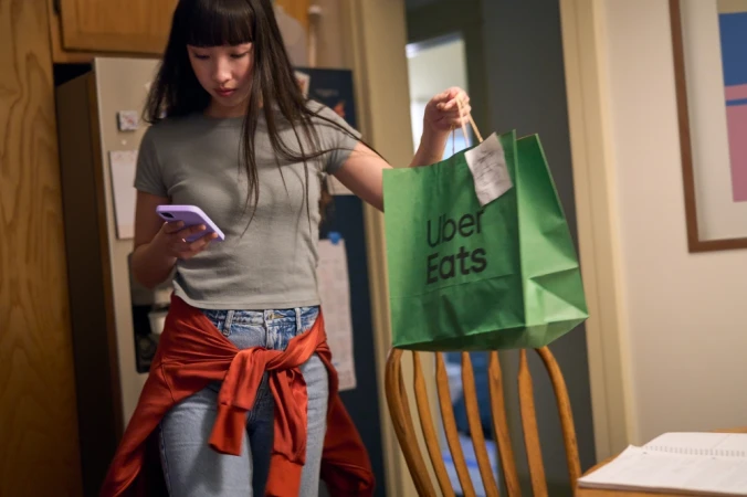 Young adult woman checks her phone while holding a green Uber Eats delivery bag inside a home with wood furniture.