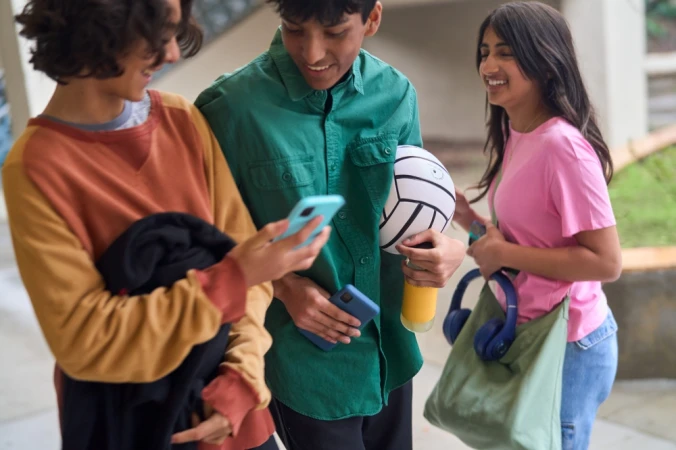 Three teenagers interact with smartphones and a plush volleyball toy in a casual indoor setting, smiling and holding personal items.