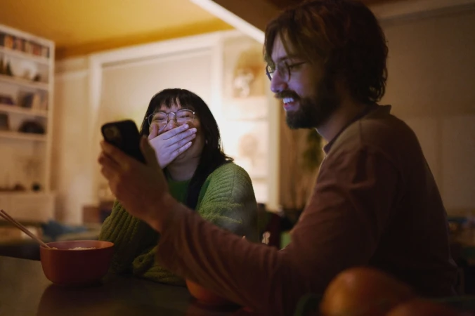 Young adults laughing at a phone screen in a warmly lit home interior, featuring a man with a beard and a woman with glasses.