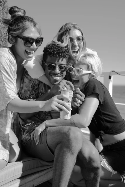 Group of four friends laughing and enjoying a beverage outdoors on a sunny day; three women and one man are together and smiling.