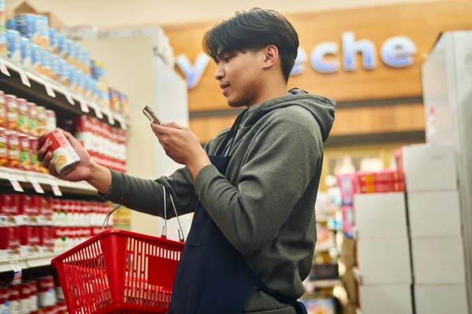 Man in apron in grocery store aisle takes photo of can of soup in hand, with a red shopping basket, next to shelves of canned food.