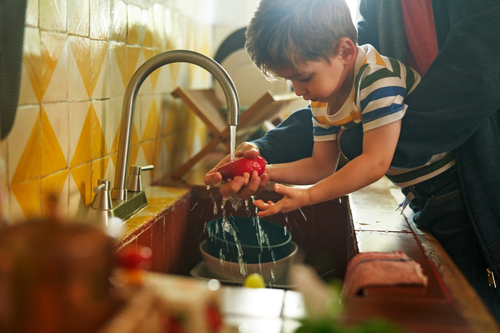 A young child washing a red apple under a kitchen faucet with water running, alongside an adult in an indoor kitchen setting.