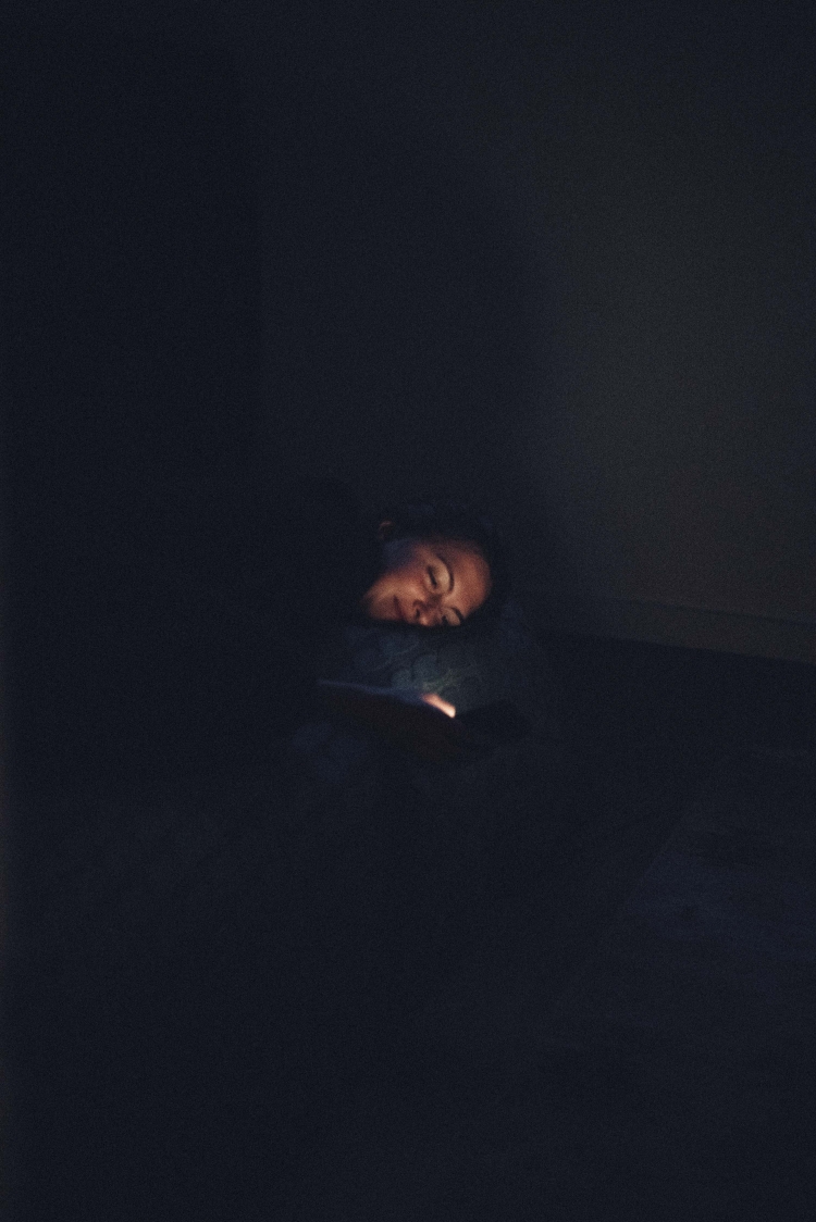 Person illuminated by phone light in dark room. Adult with head resting looks at a mobile device screen with a faint glow.