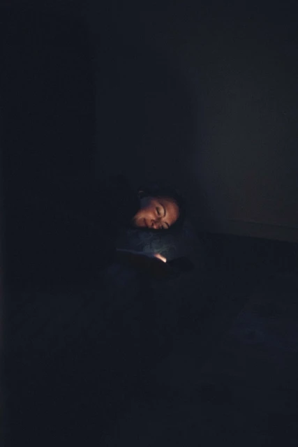 Person illuminated by phone light in dark room. Adult with head resting looks at a mobile device screen with a faint glow.