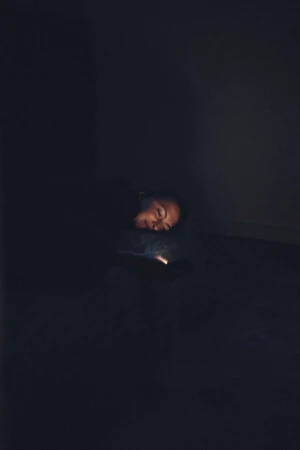 Person illuminated by phone light in dark room. Adult with head resting looks at a mobile device screen with a faint glow.