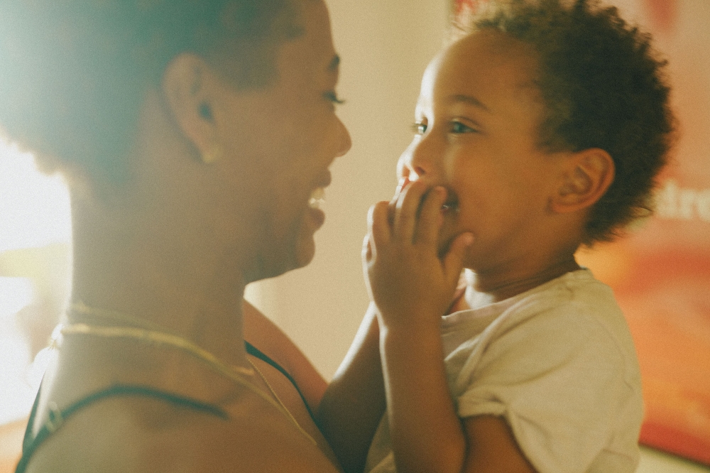 Close-up of smiling woman holding a toddler with hands up to their face indoors with an orange-red wall in the background.