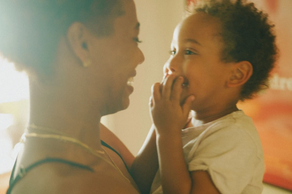 Close-up of smiling woman holding a toddler with hands up to their face indoors with an orange-red wall in the background.