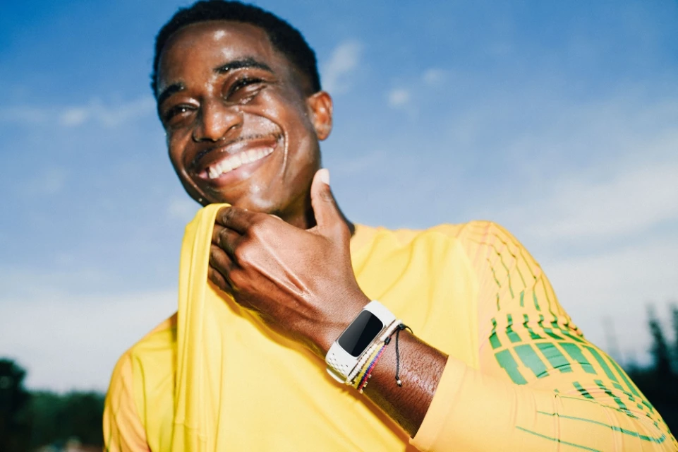 Smiling African-American man in yellow athletic shirt wipes sweat outdoors against blue sky, wearing a smart watch and colorful bracelets.