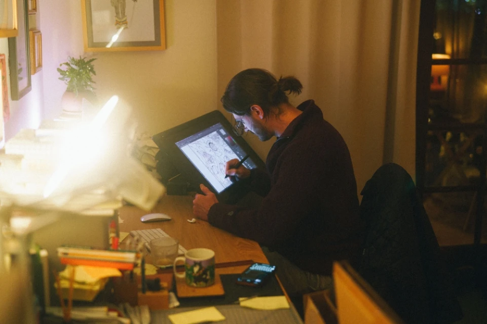 Adult male illustrator works on a digital drawing tablet in a warmly-lit home office. Artwork, a mug, and a keyboard are visible.