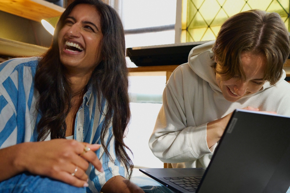 Two young adults, an East Indian woman and a Caucasian man, smile and laugh while looking at a laptop indoors in natural daylight.