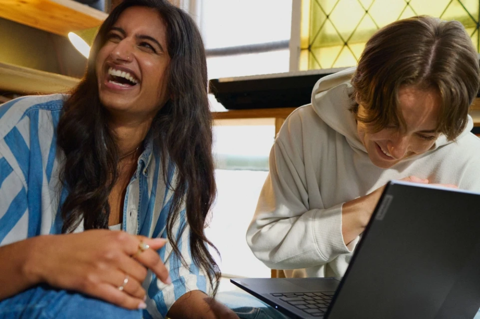 Two young adults, an East Indian woman and a Caucasian man, smile and laugh while looking at a laptop indoors in natural daylight.