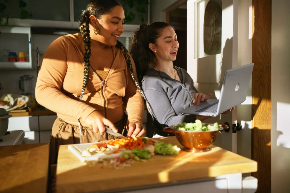 Two women in a sunlit kitchen are preparing food. One is chopping vegetables, and the other is holding a laptop, seemingly following a recipe.