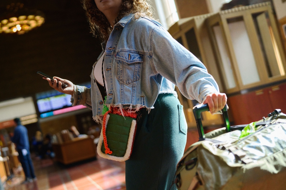 A woman in a denim jacket stands in a train station, using a smartphone while pulling a rolling suitcase. The scene is naturally lit.