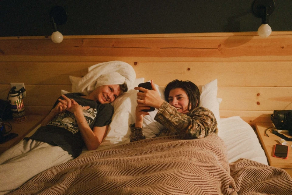 Two young adults in a bed, one with a towel on their head, looking at a smartphone in a room with wood paneled walls.