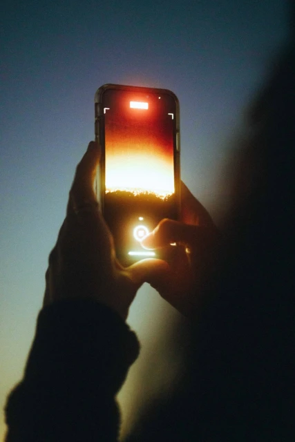 Person holds smartphone to photograph sunset or sunrise with orange glow and silhouette against a gradient blue sky in an outdoor setting.